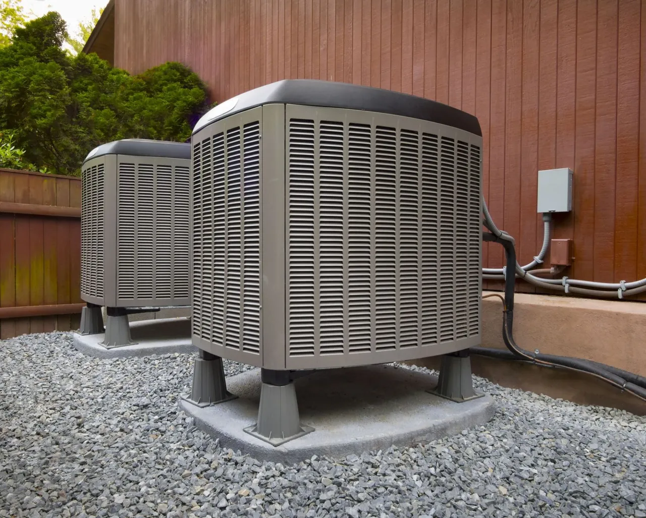 Two outdoor air conditioning units on gravel next to a brown wooden fence.
