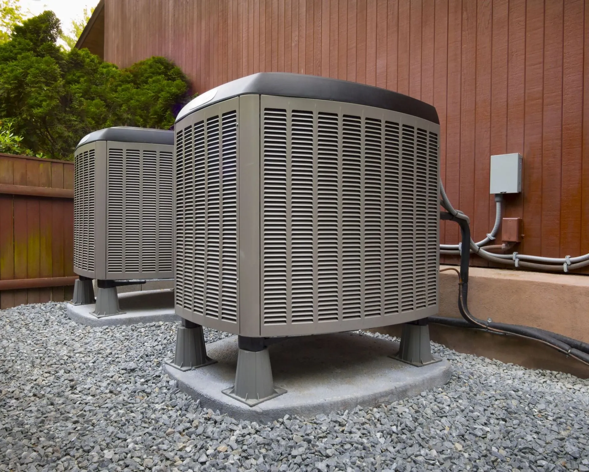 Two outdoor air conditioning units on gravel next to a brown wooden fence.